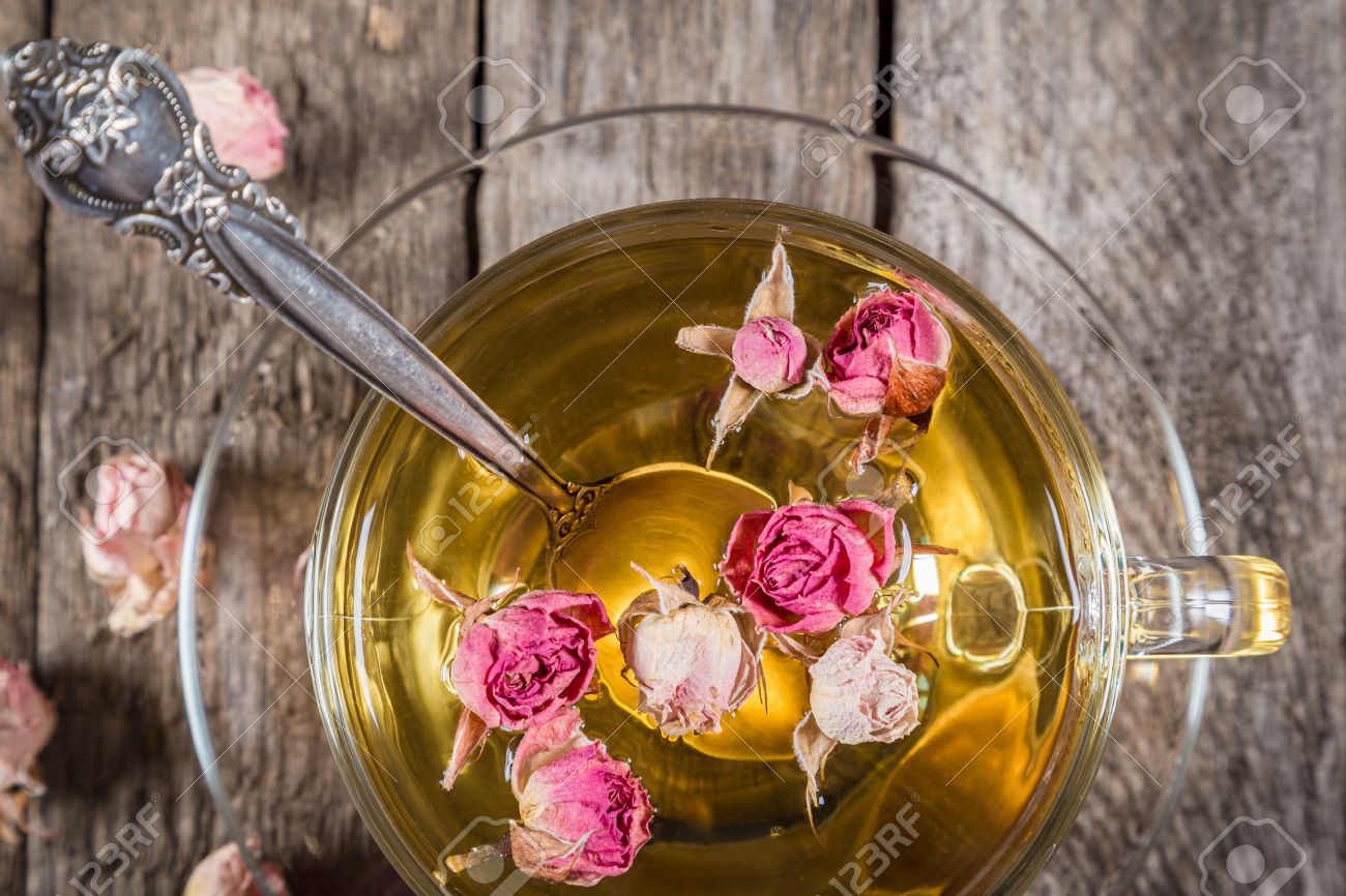 Closeup of green tea cup with teaspoon and dried rose buds on old wooden table, above view
